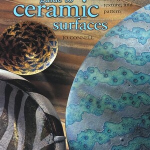 May include: The book cover for The Potter's Guide to Ceramic Surfaces by Jo Connell features a title in light blue text. It displays three ceramic objects: a small gold bowl with dark triangles, a white bowl with black stripes, and a large blue-green textured plate.