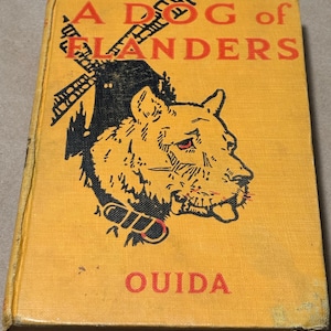 May include: A vintage hardcover book titled "A Dog of Flanders" by Ouida. The cover is a mustard yellow with black and orange illustrations of a dog and a windmill. The book's title is in orange block letters.