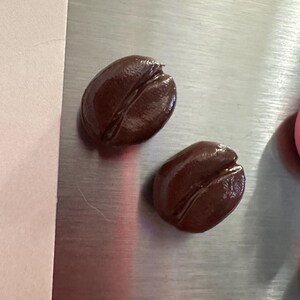 May include: Two glossy, dark brown coffee bean-shaped objects. Each bean has a prominent central groove, and the surface reflects light, suggesting a smooth texture. The beans are set against a metallic, silver background.