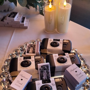 May include: A mirrored tray with a crystal-beaded edge holds three black disposable cameras wrapped in white bands labeled Help Us Capture The Memories, alongside stacks of custom matchboxes printed with Stephanie, Derek, and 07.24.2026.