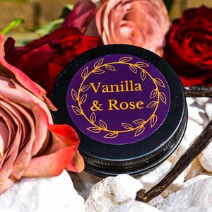 Op de afbeelding: Een klein zwart potje met een paars etiket waarop "Vanilla & Rose" staat, omringd door roze en rode rozen en vanille peulen.