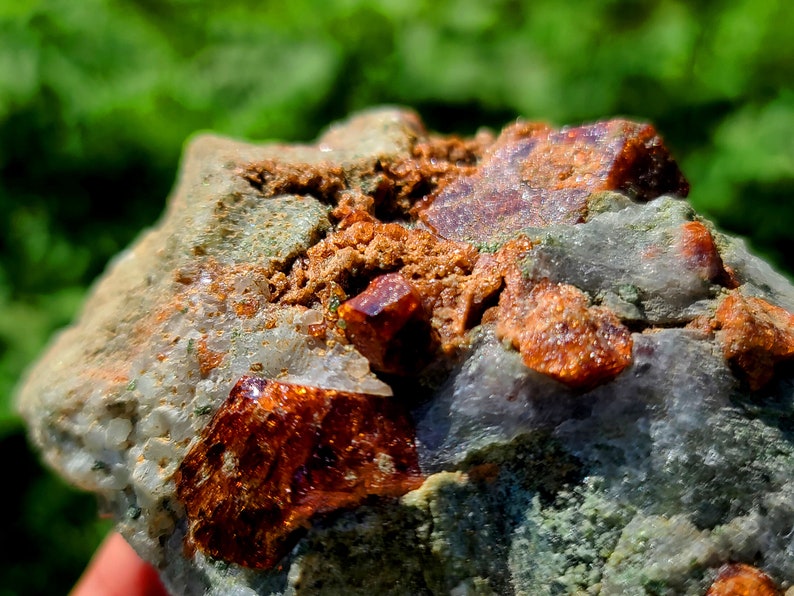 May include: A close-up of a rough, gray rock with embedded orange and red crystals. The crystals are clustered together and have a shiny, translucent appearance.