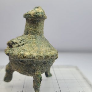 May include: A small, antique bronze incense burner with three legs. The vessel has a bulbous body, a decorative rim, and a flared top. The bronze has a weathered, green patina, suggesting age and use. The object is likely a historical artifact.