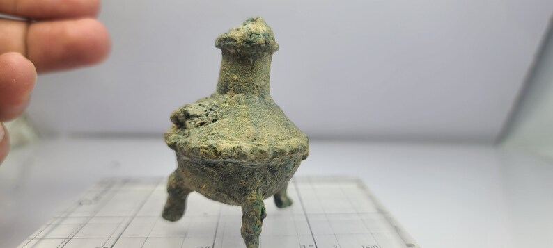 May include: A small, antique bronze incense burner with three legs. The vessel has a bulbous body, a decorative rim, and a flared top. The bronze has a weathered, green patina, suggesting age and use. The object is likely a historical artifact.