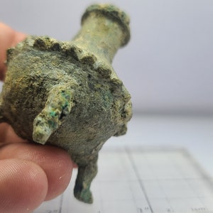 May include: A small, antique bronze vessel with a weathered, green patina. The vessel has a rounded body, three small legs, and a flared rim. The object is held in a hand.