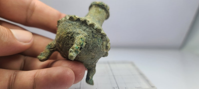 May include: A small, antique bronze vessel with a weathered, green patina. The vessel has a rounded body, three small legs, and a flared rim. The object is held in a hand.