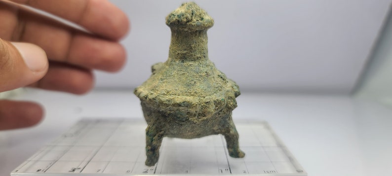 May include: A small, aged bronze incense burner with a flared base and a cylindrical top. The object has three legs and is covered in a green patina, suggesting age. The burner is set on a clear, grid-marked surface.