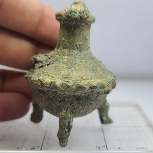 May include: A small, antique bronze incense burner with a weathered, textured surface. The vessel has a round base supported by three legs and a tall, ornate top. The color is a mix of green and brown.