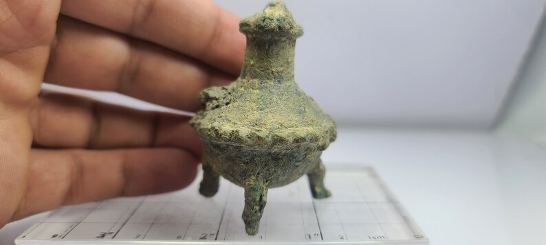 May include: A small, antique bronze incense burner with a weathered, textured surface. The vessel has a round base supported by three legs and a tall, ornate top. The color is a mix of green and brown.