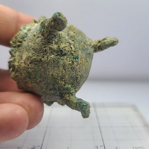 May include: A small, antique bronze vessel with a rounded body and three small, protruding legs. The surface has a textured appearance with areas of green oxidation. The vessel has a small handle on top and is held in a hand.