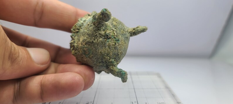 May include: A small, antique bronze vessel with a rounded body and three small, protruding legs. The surface has a textured appearance with areas of green oxidation. The vessel has a small handle on top and is held in a hand.