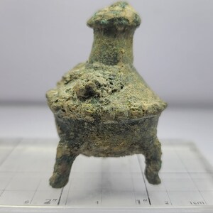 May include: A small, weathered bronze incense burner with three legs and a flared top. The vessel has a textured surface and a greenish patina, suggesting age. The burner is set against a white background.