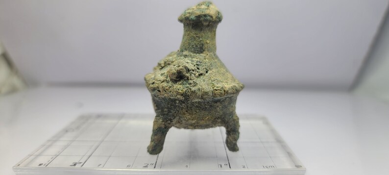 May include: A small, weathered bronze incense burner with three legs and a flared top. The vessel has a textured surface and a greenish patina, suggesting age. The burner is set against a white background.