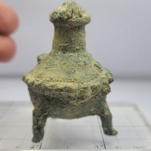 May include: A small, aged bronze incense burner with a flared base and a cylindrical top. The object has three legs and is covered in a green patina, suggesting age. The burner is set on a clear, grid-marked surface.
