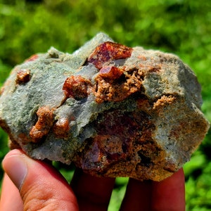 May include: A close-up of a rough, gray rock with embedded red crystals. The rock is being held in a hand.