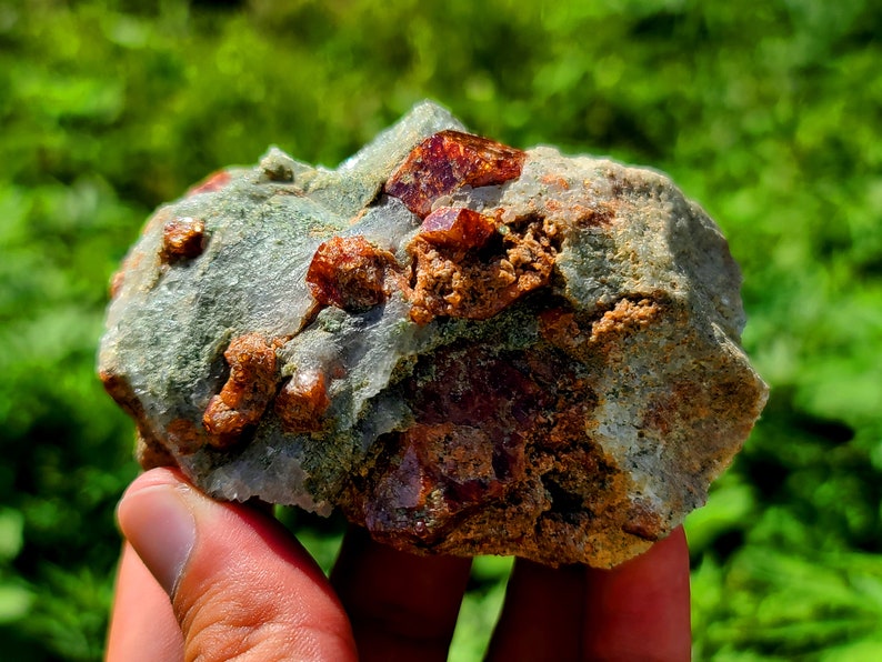 May include: A close-up of a rough, gray rock with embedded red crystals. The rock is being held in a hand.