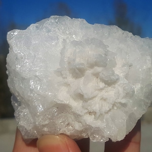Può includere: Una grande roccia bianca e cristallina con una superficie ruvida e testurizzata. La roccia ha un gruppo di cristalli più piccoli al centro.