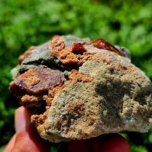 May include: A close-up of a rough, gray rock with embedded red crystals. The rock is being held in a hand.