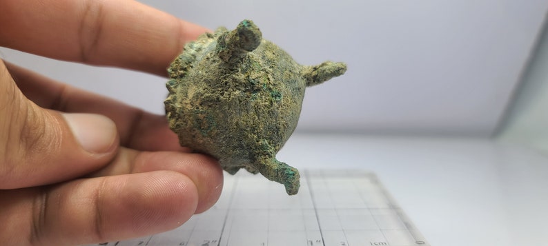 May include: A small, antique bronze vessel with a rounded body and three small, protruding legs. The surface has a textured appearance with areas of green oxidation. The vessel is held in a hand, with a ruler in the background.