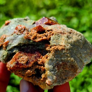 May include: A rough, gray rock with embedded red and orange crystals. The rock is being held in a hand.