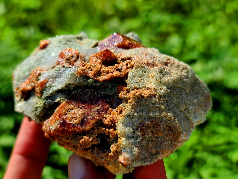 May include: A rough, gray rock with embedded red and orange crystals. The rock is being held in a hand.