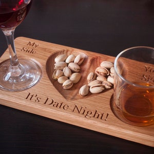 May include: Wooden serving tray with two indented spaces for glasses, a heart-shaped space for snacks, and the text "It's Date Night!"  The tray has the words "My Side" and "Your Side" engraved on either side of the heart-shaped space.  A glass of red wine and a glass of whisky are on the tray, along with pistachios in the heart-shaped space.