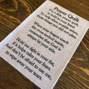 May include: A small, rectangular prayer quilt with black text on a light beige fabric. The text includes a poem about faith and comfort. The quilt is designed to be held and offers solace during difficult times.