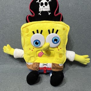 May include: A plush toy of Spongebob Squarepants dressed as a pirate. The character is bright yellow with large blue eyes, a pink tongue sticking out, and a black pirate hat featuring a white skull and crossbones. It wears a brown vest.