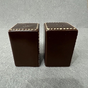 Op de afbeelding: Een paar vintage bruine zout- en peperstrooiers. Elke strooier is een rechthoekig prisma met een licht taps toelopende vorm. De bovenkant van elke strooier heeft drie kleine gaatjes. De randen zijn versierd met een stippenpatroon.