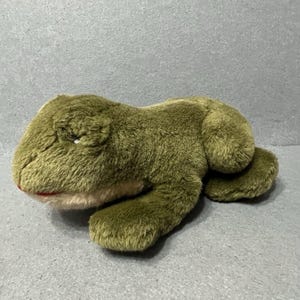 Op de afbeelding: Een groene en beige pluche kikker. Het speelgoed heeft een zacht, getextureerd uiterlijk met een rode mond en kleine, ronde ogen. De kikker ligt op een grijze ondergrond.