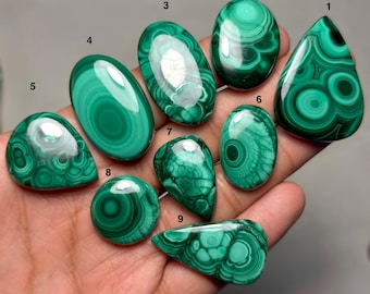Pode incluir: Nove cabochões de malaquita polidos em várias formas, incluindo oval, gota e redonda, cada um exibindo padrões distintos de faixas concêntricas verdes. As pedras são numeradas de um a nove, mostrando variações naturais em tons de verde escuro.