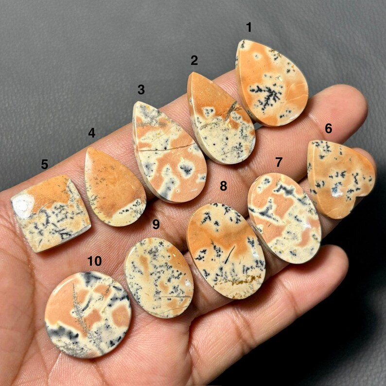 May include: A collection of ten polished gemstones in various shapes, including teardrop, oval, and square. The stones feature a cream base with orange and black dendritic patterns. The gemstones are held in a hand.