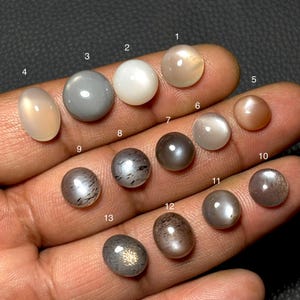 Może przedstawiać: Kolekcja trzynastu wypolerowanych kamieni szlachetnych o różnych kształtach i kolorach, w tym brzoskwiniowym, szarym, białym i brązowym. Kamienie szlachetne są prezentowane na dłoni, prezentując ich różne tekstury i połysk. Kamienie szlachetne są ponumerowane od 1 do 13.