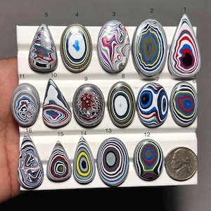 Könnte beinhalten: Eine Sammlung von sechzehn polierten Steinen mit einzigartigen, farbenfrohen Mustern. Die Steine haben verschiedene Formen, darunter oval und tropfenförmig, und weisen konzentrische Designs in Rot-, Blau-, Weiß- und Schwarztönen auf. Eine Münze dient als Größenvergleich.