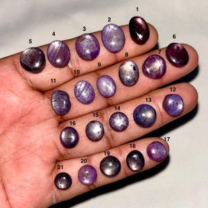 May include: A collection of twenty-one oval and round cabochon-cut star sapphire gemstones in various shades of purple and deep violet. Each stone is individually numbered from one to twenty-one, displaying unique internal light reflections and patterns.
