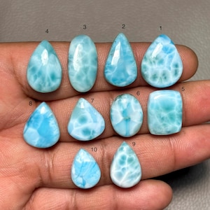 May include: A collection of ten polished larimar gemstones in various shapes, including teardrop, oval, and rectangular. The stones display a range of light blue hues with white marbling, creating a sea-like appearance. The gemstones are held in a hand.