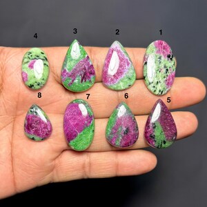 Könnte beinhalten: Acht polierte Rubin-Zoisit-Edelsteine in verschiedenen Formen, darunter oval und tropfenförmig. Die Steine weisen eine Mischung aus leuchtendem Grün und tiefem Magenta auf, einige zeigen schwarze Sprenkel.