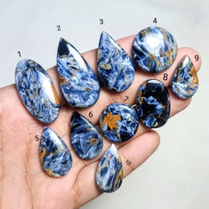 May include: A collection of ten polished Pietersite gemstones in various shapes, including oval, teardrop, and round. The stones display a striking mix of blue, gold, and black colors, with swirling patterns and a glossy finish. The gemstones are held in a hand.