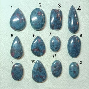 May include: A collection of twelve polished gemstones in various shapes, including teardrop, oval, and rectangular. The stones have a blue-gray base color with red inclusions, creating a speckled appearance. The gemstones are displayed on a light-colored surface.