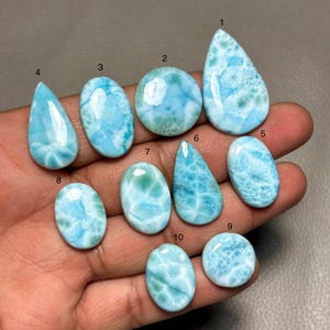 Puede incluir: Una colección de diez piedras preciosas de larimar pulidas en varias formas, incluyendo lágrima, ovalada y redonda. Las piedras muestran una gama de patrones azules y blancos, algunos con apariencia veteada. Las piedras preciosas se sostienen en una mano.