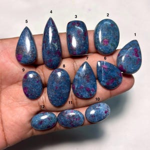 May include: Twelve polished cabochon gemstones featuring a deep blue base color with scattered magenta and pink inclusions. Each stone is numbered from one to twelve and displays a smooth, rounded surface with varying shapes including teardrop, oval, and rectangular forms.