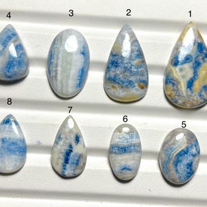 May include: Eight polished gemstones with blue, white, and yellow patterns. The stones vary in shape, including teardrop and oval. The gemstones are displayed on a white surface with a grid pattern.