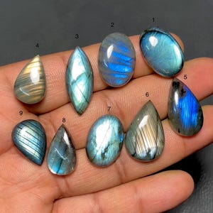 May include: A collection of nine polished labradorite gemstones in various shapes, including oval, teardrop, and marquise. The stones display iridescent flashes of blue, green, and gold, with visible internal patterns. The gemstones are held in a hand.