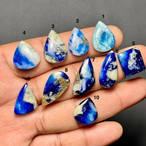 May include: A collection of ten polished gemstones in various shapes, including teardrops, ovals, and a square. The stones display a mix of blue, white, and gold colors, with unique patterns and textures. The gemstones are held in a hand.