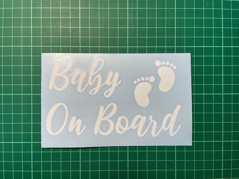 May include: White vinyl sticker with the text "Baby On Board" in a cursive font. Two white footprints are to the right of the text.