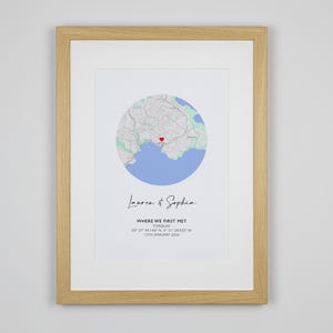May include: A framed print featuring a circular map of Torquay with a red heart icon. Below the map, the text reads Lauren & Sophia, WHERE WE FIRST MET, TORQUAY, 50 27 44.146 N, 3 31 28.705 W, 13TH JANUARY 2024. The frame is light-colored wood.