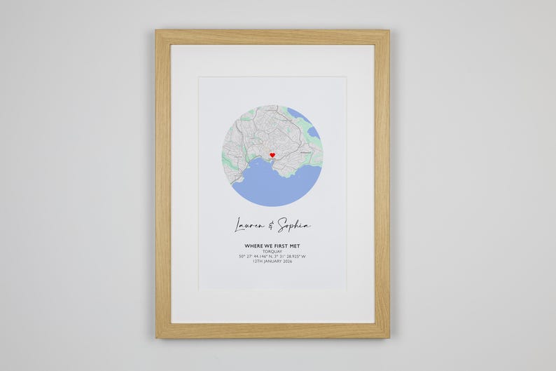 May include: A framed print featuring a circular map of Torquay with a red heart icon. Below the map, the text reads Lauren & Sophia, WHERE WE FIRST MET, TORQUAY, 50 27 44.146 N, 3 31 28.705 W, 13TH JANUARY 2024. The frame is light-colored wood.