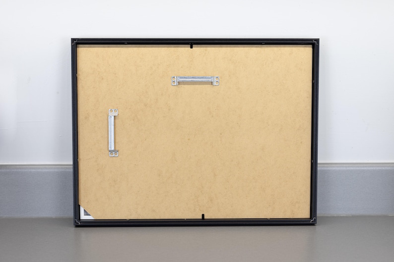 May include: Back of a framed picture with a black frame. The backing is a light brown color. Metal hanging hardware is attached to the back. The frame is resting against a white wall and a gray floor.