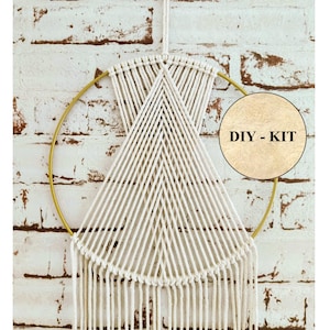 Könnte beinhalten: Ein weißer Makramee-Wandbehang mit einem goldenen Reifen, der an einer weißen Backsteinwand hängt. Der Makramee hat ein geometrisches Muster mit Fransen am unteren Rand. Ein brauner Kreis mit dem Text "DIY - KIT" befindet sich in der oberen rechten Ecke des Bildes.