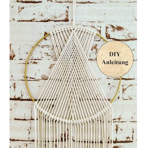 Könnte beinhalten: Ein weißer Makramee-Wandbehang mit einem goldenen Reifen und langen weißen Fransen. Der Makramee ist in einem geometrischen Muster. Ein Holzschild mit dem Text "DIY Anleitung" befindet sich im Hintergrund.